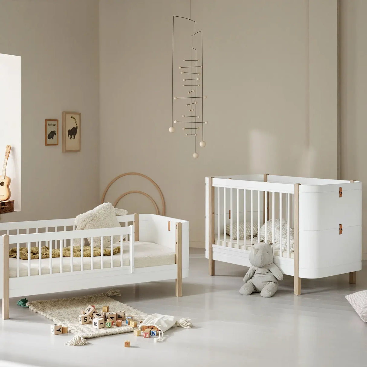 A beautifully styled nursery featuring an Oliver Furniture cot and junior bed in a Scandinavian-inspired design. The furniture is crafted from white and natural wood, with soft neutral tones creating a calming atmosphere. The cot has a modern aesthetic with white slats and leather detailing, while the junior bed features a similar design with a half-railed frame. The room is decorated with a plush teddy bear, wooden alphabet blocks, framed animal prints, and a delicate mobile, adding a warm touch