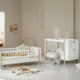 A beautifully styled nursery featuring an Oliver Furniture cot and junior bed in a Scandinavian-inspired design. The furniture is crafted from white and natural wood, with soft neutral tones creating a calming atmosphere. The cot has a modern aesthetic with white slats and leather detailing, while the junior bed features a similar design with a half-railed frame. The room is decorated with a plush teddy bear, wooden alphabet blocks, framed animal prints, and a delicate mobile, adding a warm touch