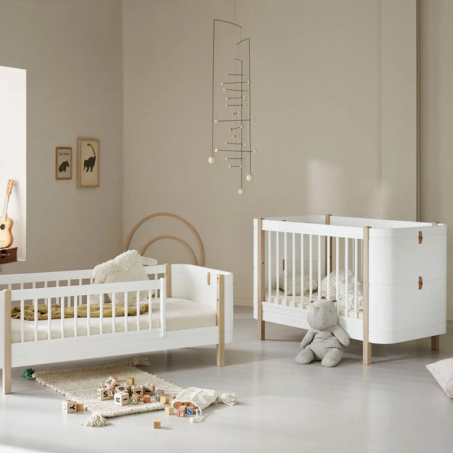 A beautifully styled nursery featuring an Oliver Furniture cot and junior bed in a Scandinavian-inspired design. The furniture is crafted from white and natural wood, with soft neutral tones creating a calming atmosphere. The cot has a modern aesthetic with white slats and leather detailing, while the junior bed features a similar design with a half-railed frame. The room is decorated with a plush teddy bear, wooden alphabet blocks, framed animal prints, and a delicate mobile, adding a warm touch