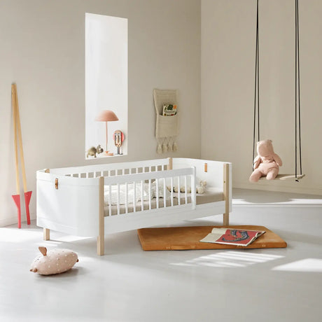A serene and stylish children's room featuring the Oliver Furniture Wood Mini+ Junior Bed in a Scandinavian-inspired design. The bed has a sleek white finish with natural oak legs and leather detailing, creating a modern yet timeless look. The room is softly lit with natural sunlight, highlighting minimalistic decor elements, including a wooden swing with a plush teddy bear, a cozy floor cushion, and a reading nook with books on a leather mat.